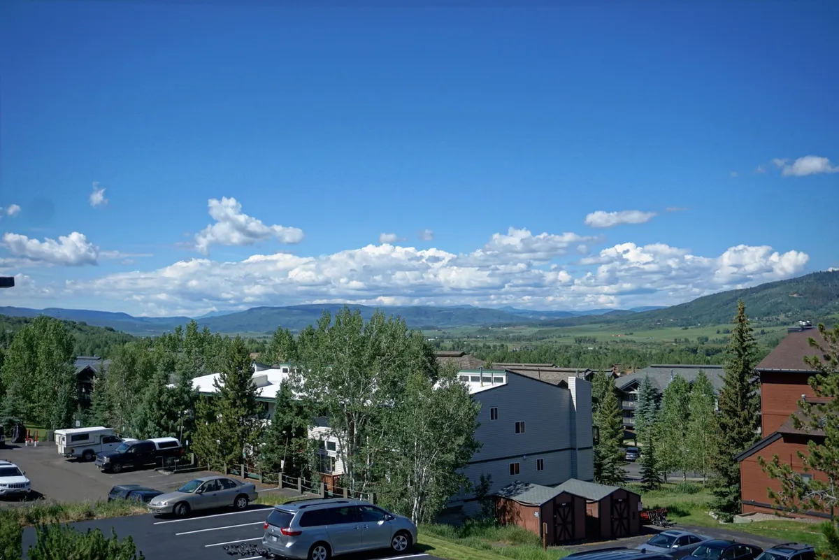 A panoramic view of a mountain range in the background with clouds overhead, green trees in the foreground, and buildings and parked cars visible. The scene suggests proximity to nature trails and the inviting landscape surrounding the property.