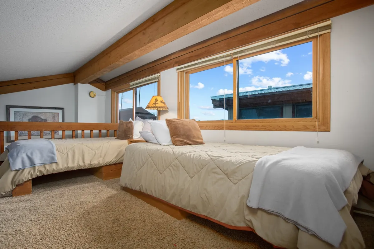 A unique loft area features exposed wooden beams, large windows framing mountain views, and cozy bedding. The space exudes warmth and adventure, hinting at a special hideaway perfect for older kids or adults. The view out the window adds to the magical atmosphere.