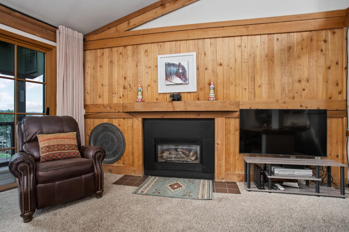 A warm, inviting corner of the condo featuring a glowing fireplace, comfortable armchair, wooden wall, and tasteful artwork. The scene offers an intimate setting, ideal for cozy evenings and intimate conversations.