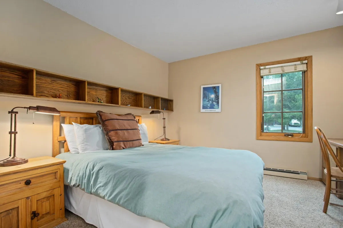 This image shows a cozy bedroom with a soft turquoise cover on the bed, wooden nightstands, rustic shelving, and a window that allows natural light to flood the space. The room feels peaceful and inviting, ideal for rest and relaxation after a day of adventure.