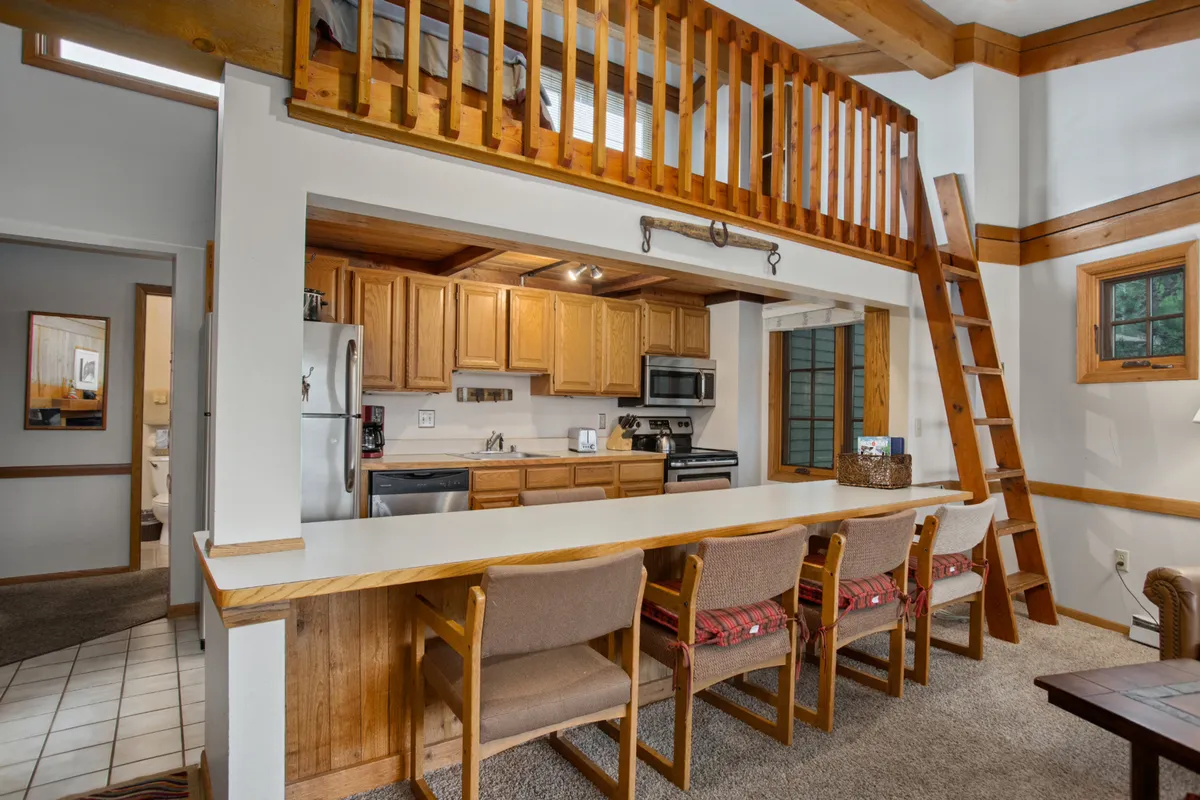 A cozy kitchen scene with a prominent island, a wooden ladder leading to an open loft space, and expansive windows that flood the area with morning sunlight. The layout encourages group cooking, socializing, and relaxation, with ample counters and a warm, communal vibe.