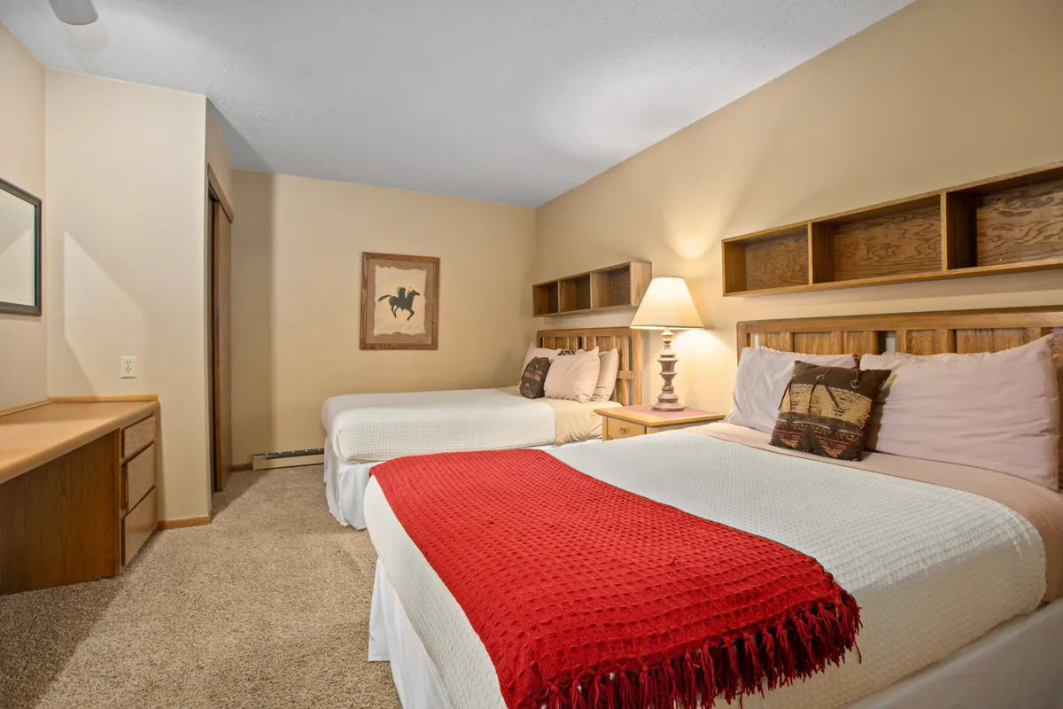 A bright room with two comfortable beds, a decorative red throw, and a lamp create an inviting atmosphere perfect for families or friends. The arrangement suggests ample space for guests to relax together or enjoy some quiet time.