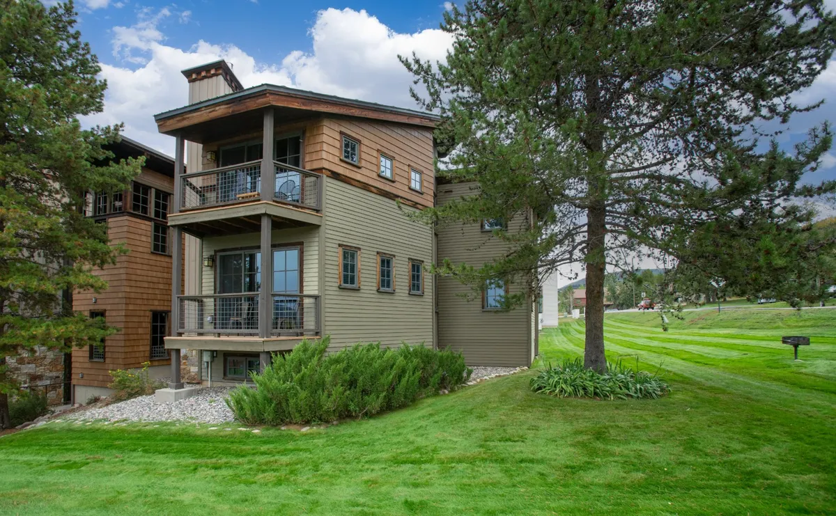 The image showcases the condo’s inviting wooden balcony overlooking a lush green lawn, framed by a majestic tall pine tree. Large windows catch the sunlight and hint at bright, cozy interiors, while the expansive yard below offers plenty of space for family gatherings or outdoor relaxation. The overall scene is warm and welcoming, evoking the spirit of mountain escapes in Steamboat Springs.