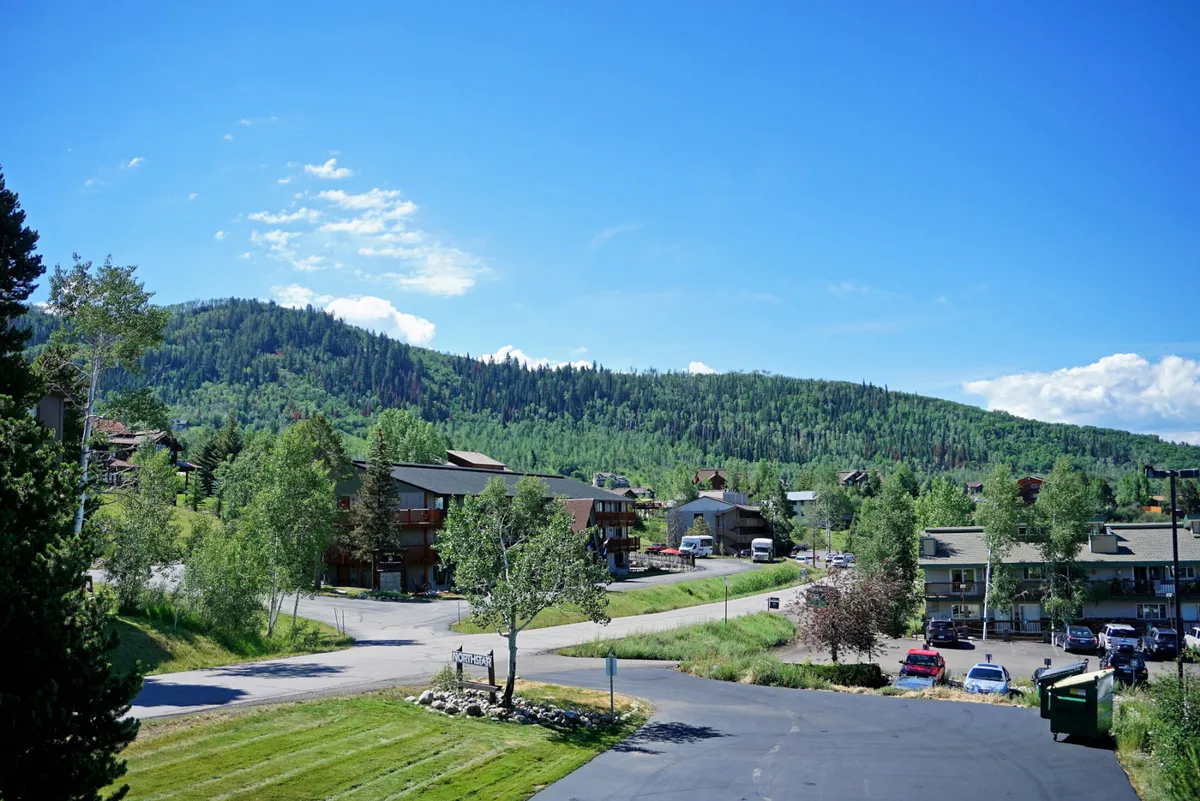 A sweeping view of mountains and lush greenery, with buildings and a winding road in the foreground. The scene captures the relaxed yet adventurous spirit of exploring nature, hiking, biking, and enjoying local events.