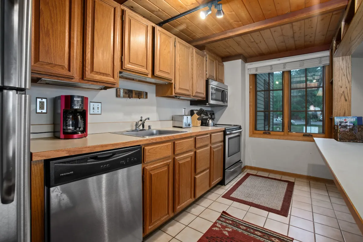 A well-appointed kitchen illuminated by natural light from a large window features wooden cabinetry, stainless steel appliances, and a welcoming coffee maker station. The space is inviting and practical for everything from casual breakfasts to festive dinners.