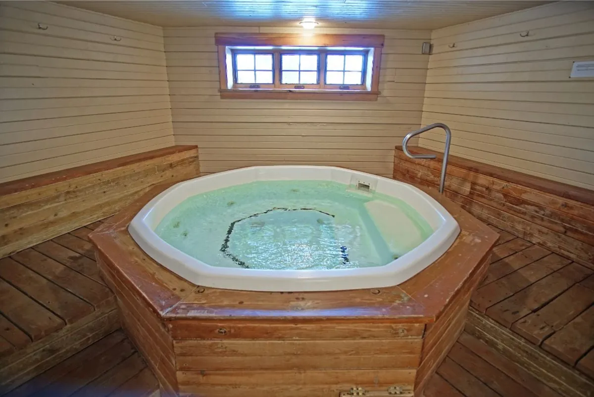 A tranquil community wellness area featuring a jacuzzi surrounded by wooden seating and large windows letting in natural light. The scene evokes spa-like relaxation and is clearly designed for group or solo unwinding after outdoor activities.