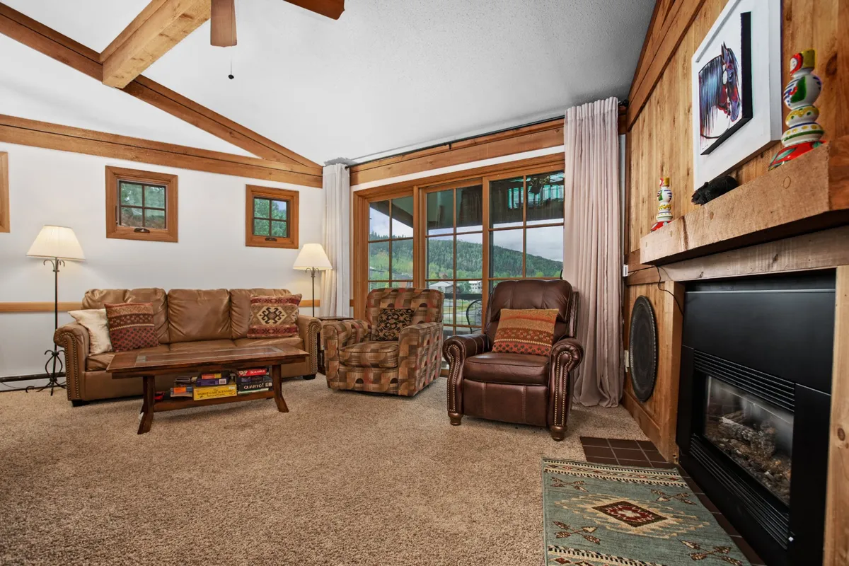 A welcoming living room scene with a fireplace as the focal point, flanked by large windows offering views and natural light. The space includes a comfortable couch and armchairs arranged for conversation or relaxation.