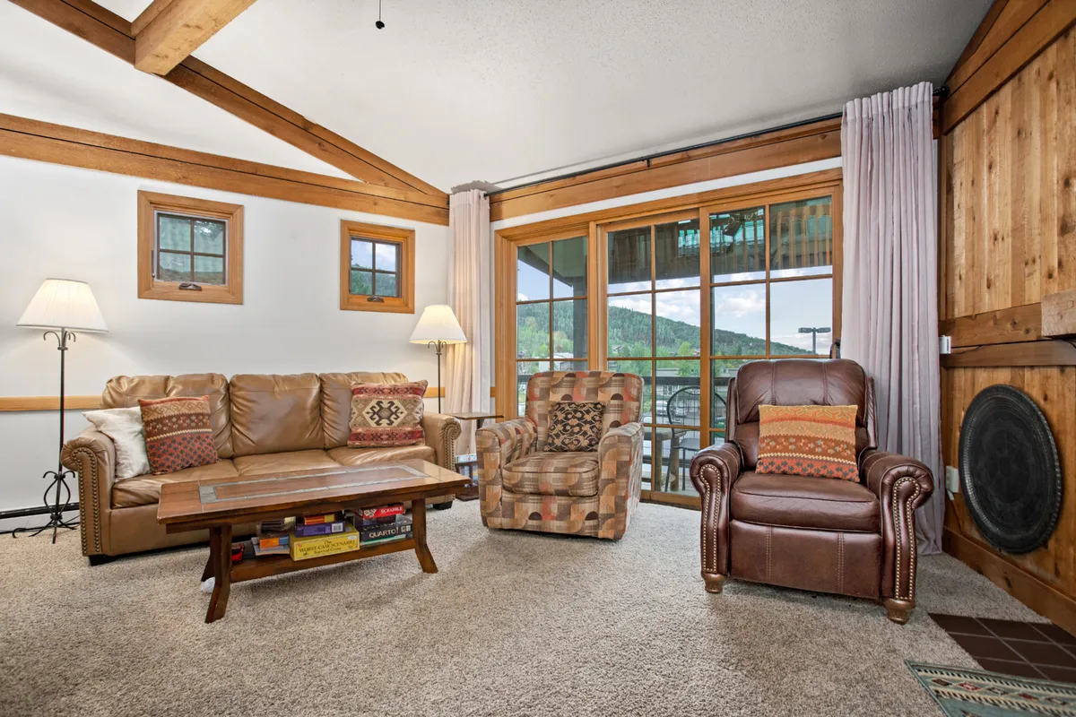 A cozy living room interior featuring a sofa, armchair, coffee table, and large windows that frame the mountainous outdoors. The space is bathed in natural light, inviting socialization and relaxation while offering glimpses of the sunset.