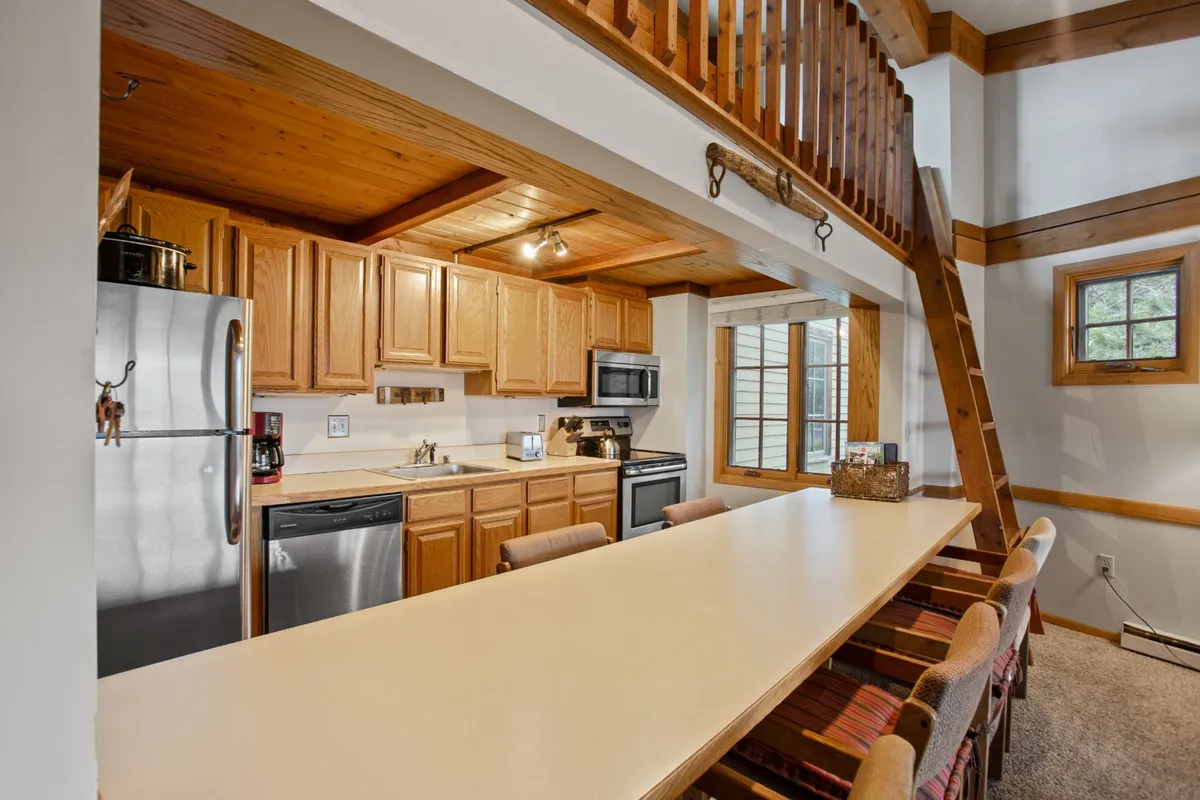 A spacious, communal kitchen features a large island with seating for several people, stainless steel appliances, and warm wooden cabinetry. The open layout includes a lofted area and windows that let in natural light, creating an inviting space ideal for both cooking and socializing.