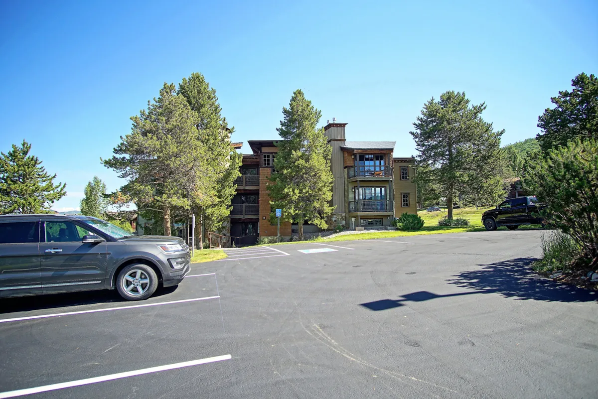 Shows the building facade nestled among tall, leafy trees, with a view of the parking area and natural surroundings. The relaxed mood suggests a welcoming environment for extended stays, convenient access, and enjoyable strolls through lush grounds.